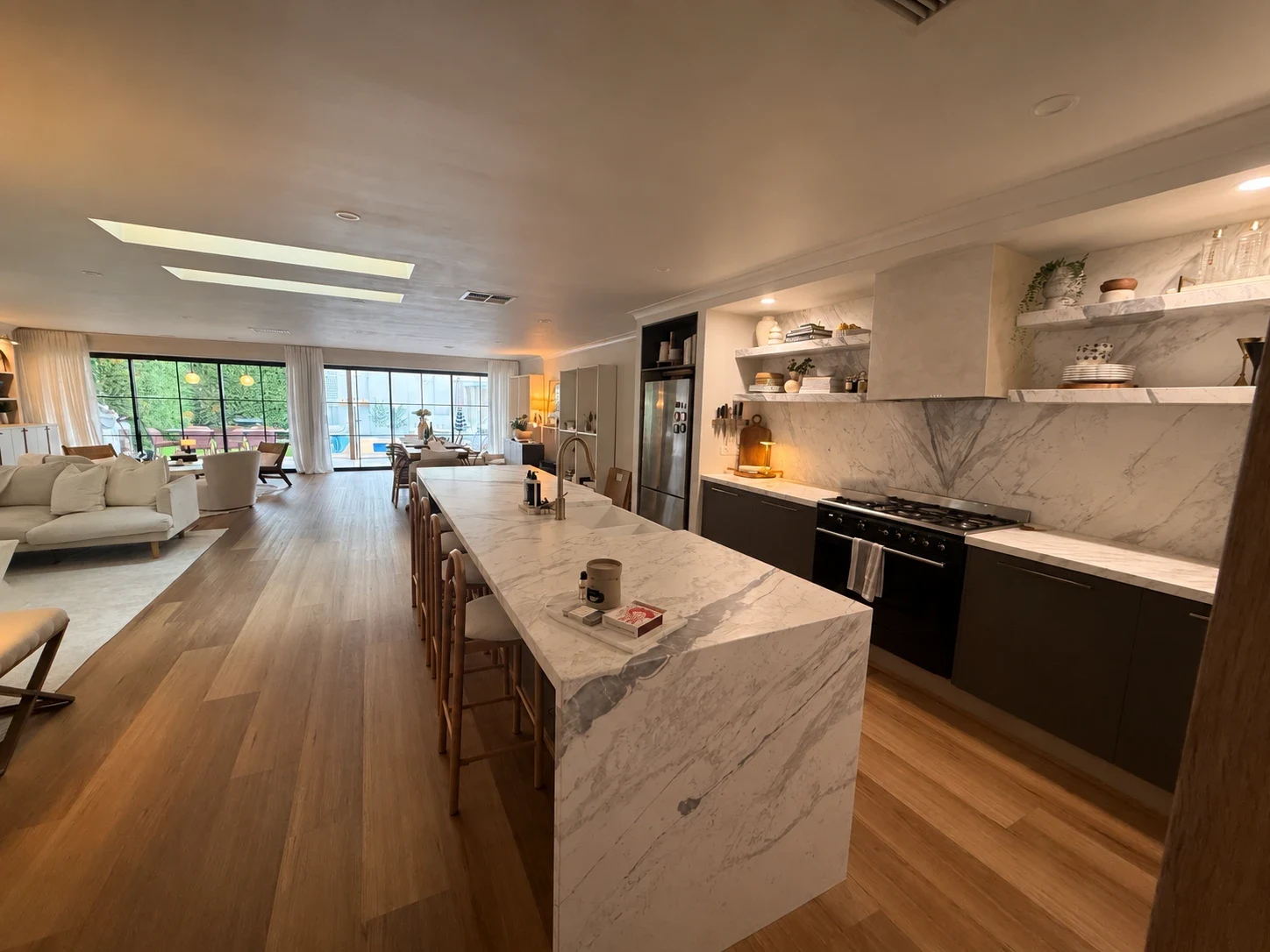 A long open-plan kitchen, living and dining room with a marble waterfall island, marble splashback, timber floors and black-framed doors onto the garden