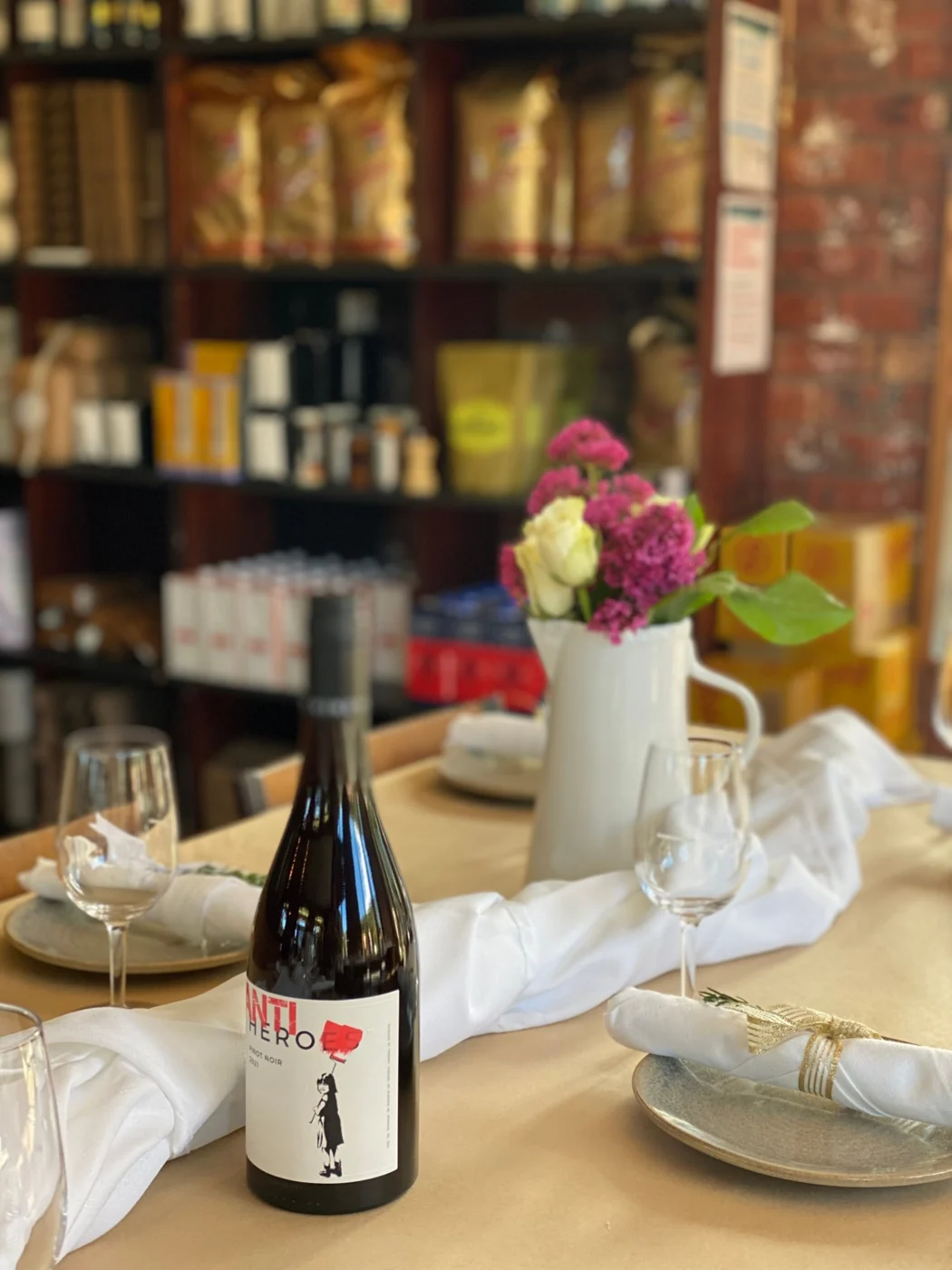 A relaxed afternoon table in a deli: white linen runner, a jug of pink and white flowers, wine glasses and napkins tied with gold ribbon and rosemary